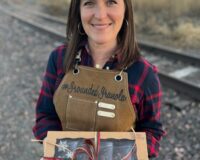 A woman smiling at the camera while holding a holiday box of assorted granola wrapped with red and green twine.