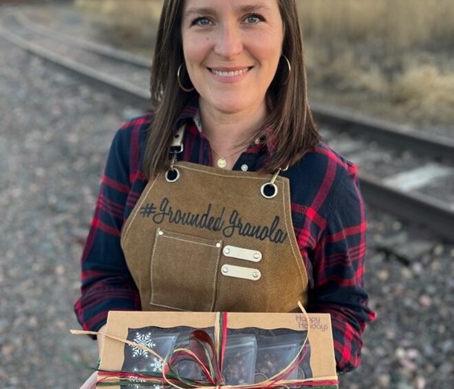 A woman smiling at the camera while holding a holiday box of assorted granola wrapped with red and green twine.